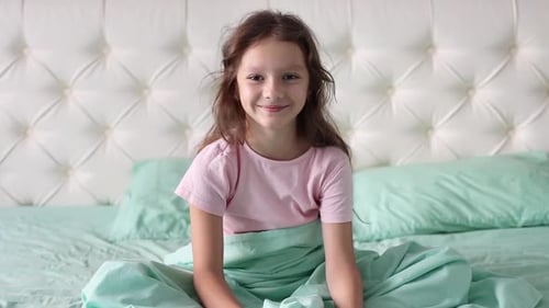 A little girl sits on a bed in pajamas and smiles at the camera, and than falls on the bed