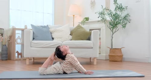 Una hija preescolar asiática practica una postura flexible doblada hacia atrás en una colchoneta de yoga en casa, imitando el bienestar