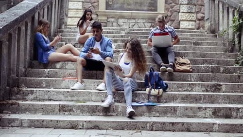 Teenage students with gadgets enjoying a sunny summer day on university campus steps