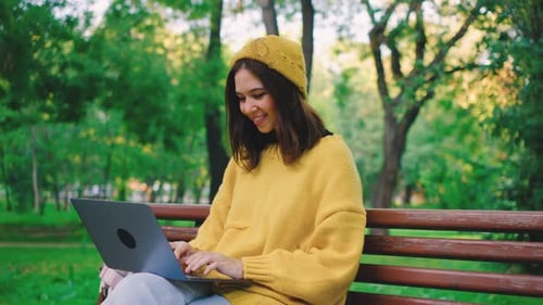 A Joyful Woman is Sitting on a Bench in the Park Happily Working on Her Laptop While Embracing the