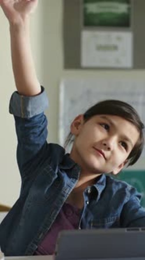Vertical video: Pausing, student in denim shirt raising hand in class to answer teacher, tablet