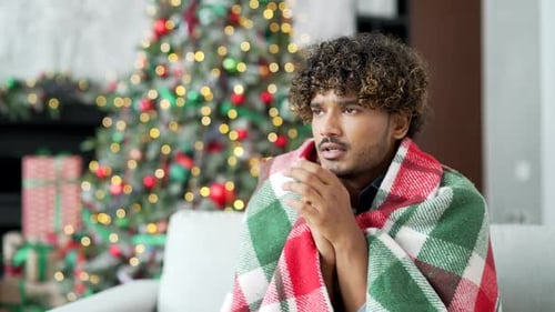 A young handsome man covered with a blanket froze sitting on sofa in living room at home during