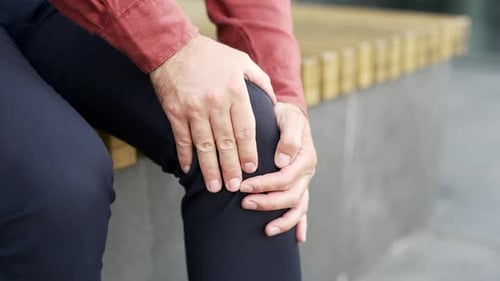 Male sitting outdoors on bench in city, hands on knee indicating discomfort or injury.