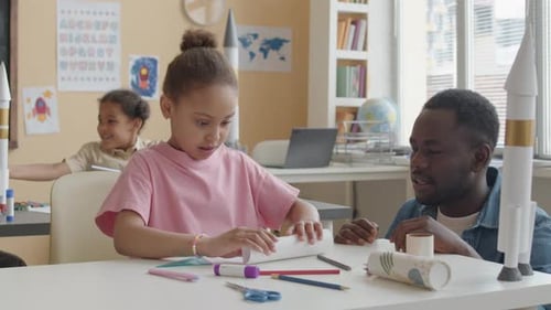 Girl Creating Paper Rocket in Classroom with Teacher