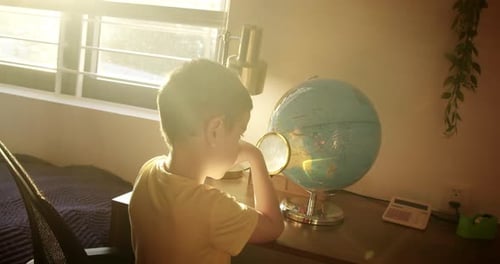 Young Boy Studies Globe with Magnifying Glass at Desk