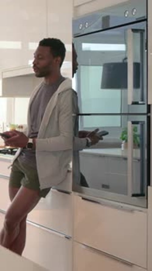 Vidéo verticale : Un homme penché en sweat à capuche fait défiler un smartphone près d'un four encastré dans une cuisine lumineuse