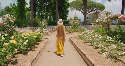 Woman walks down a garden path lined with blooming roses, her long hair flowing behind her.