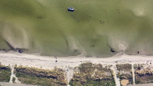 An aerial view of a lone boat anchored near a sandy beach, with clear shallow water revealing the se