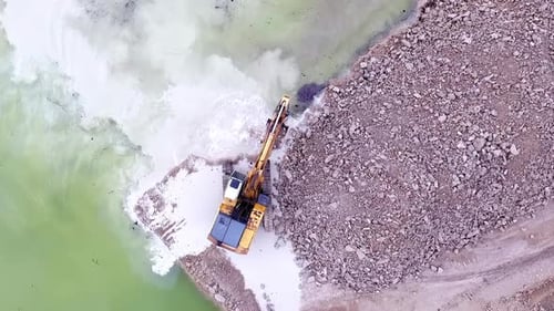 Drohnenaufnahme eines Baggers von oben nach unten, der an einer felsigen Küste neben türkisfarbenem Wasser arbeitet, schwer