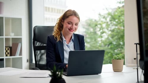 in a Modern Office with a Large Window a Beautiful Worker in a Blue Shirt with a Laptop Looks at the