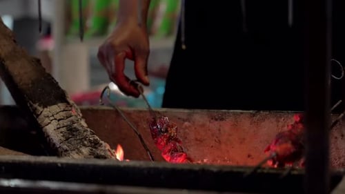 A man’s hand turning meat kabob skewers on an open flame grill in the outdoors.