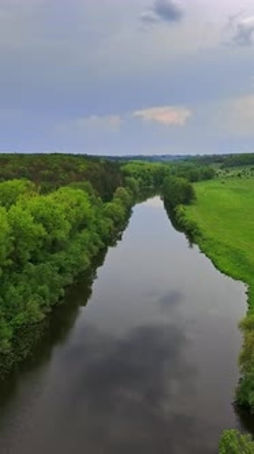 Narrow river with meadow on one side and forest on another. Cloudy sky reflecting in the water.