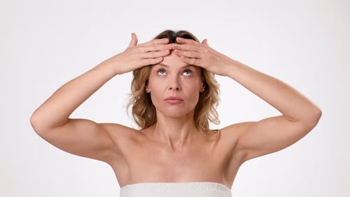 Woman Touching Her Forehead While Standing Against a Plain Background in a Studio Setting