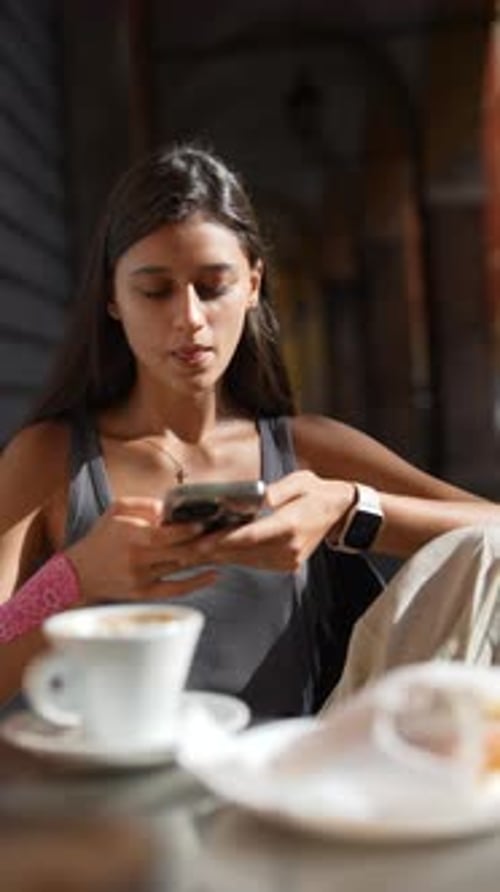 A Young Woman Enjoys a Coffee Shop Ambiance Using Her Mobile Device and Sipping a Drink