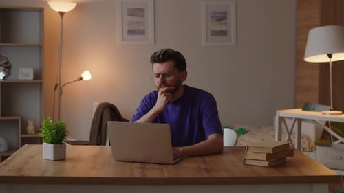 A Man in a Purple Tshirt Sits at a Table in His Room and Works on a Laptop A Man Sitting at a