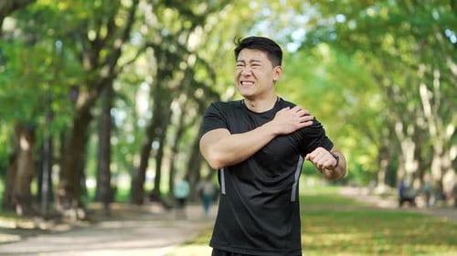 Adult asian athletic man experiencing acute shoulder pain during morning exercise in urban city park