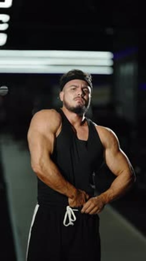 Muscular Athlete in a Black Tank Top Poses Under Bright Gym Lights Flexing Powerful Arms and Core