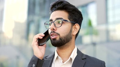 Smiling bearded businessman in formal suit talking on smartphone standing on street near office
