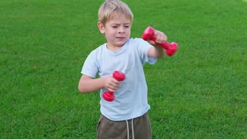 Sporty Little Boy Lifting Dumbbells on a Green Lawn Joyful Toddler Doing Exercise Against Nature