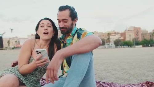 Romantic couple uses mobile phone while sitting on the beach on buildings background