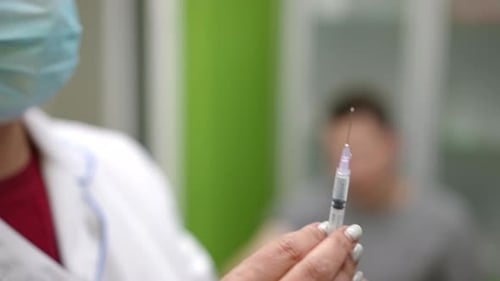 A Nurse in a White Coat and a Mask Prepares a Syringe with Medicine for Injection