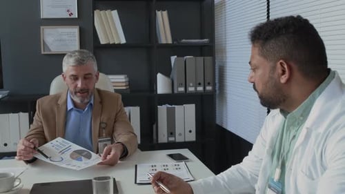 Clinical Manager Presenting Report while DoctorTaking Notes at Meeting in Office