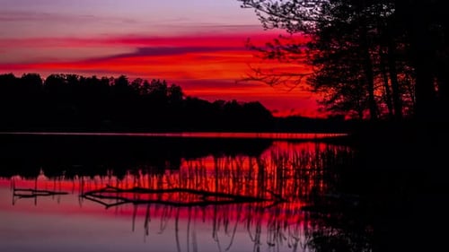 Statische Ansicht des roten Himmels bei Sonnenuntergang über Wasser im Zeitraffer, mit Bäumen am Ufer des Sees in der