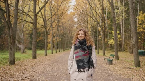 Woman with Curly Hair and Scarf Walks Through an Autumn Park Surrounded By Colorful Trees and Fallen