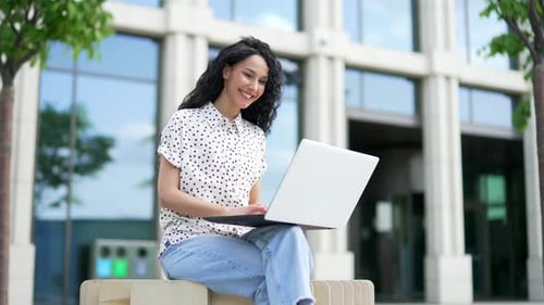 Young female student works typing on a laptop sitting on a bench in the campus space near