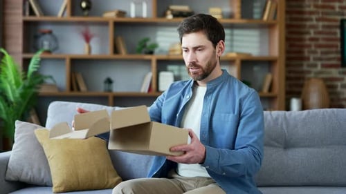 Unhappy man received a parcel with wrong product sitting on sofa in living room at home looking