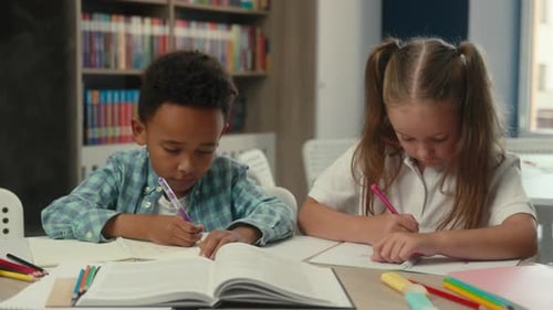 Zwei konzentrierte Kinder, multiethnische Kleinkinder, die gemeinsam in der Schulbibliothek lernen, Schüler schreiben