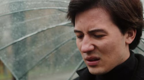 Jeune homme dans un manteau debout sous un parapluie transparent en gros plan et se mouchant le nez dans un