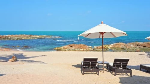 Sun umbrella and deckchair on white empty beach with sea in background. Slow-motion