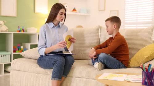 Professioneller Psychologe zeigt einem kleinen Jungen auf dem Sofa drinnen Bilder von traurigen und glücklichen Gesichtern