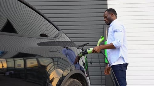 Black Man Plugging in Charging Cable to Electric Vehicle and Charges Batteries at Station