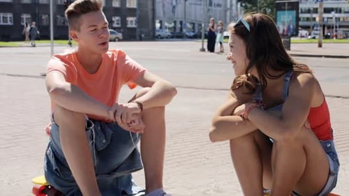 Smiling teenage couple chatting with penny boards on a sunny city street vacation