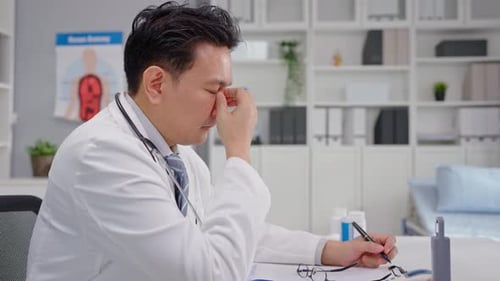 Asian stressed male doctor sitting on working table in hospital office.