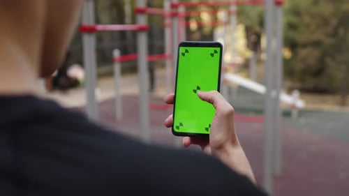 Sportsman Clicking and Swiping Smartphone with a Mockup Green Screen Standing on the Outdoor Workout