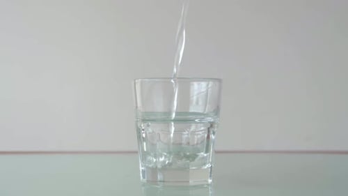Wasser in kurzes Glas auf dem Tisch mit Reflexion gießen, immer noch Nahaufnahme in minimalistischem Setup