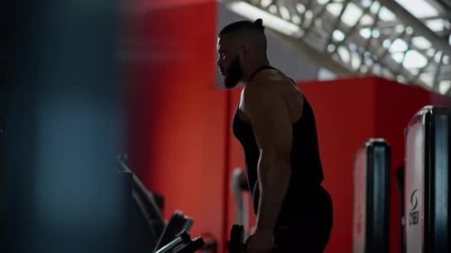 Muscular Bearded Man in Black Tank Top Performing Bicep Curl with Dumbbell Silhouetted Against Gym