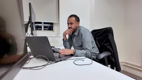 South Asian Man Working On Laptop Computer At Office Desk With Mouse Pad And Second Monitor For