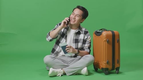 Full Body Of Asian Male Traveler With Luggage And Passport Talking On Smartphone In The Green Screen