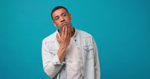 Pensive Young African American Man Thinking and Touching Chin Standing Over on Blue Studio
