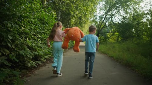 Back View of Siblings Walking on Path While Girl Lifts Teddy Bear