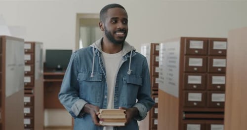 Dolly Shot of Carefree African American Man Holding Books Walking in Library Hall Looking at Shelves
