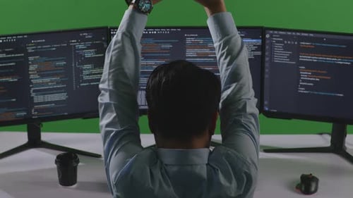 Müder asiatischer Mann dehnt sich beim Schreiben von Code mit mehreren Computerbildschirmen auf grünem Bildschirm