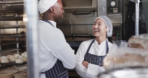 Happy diverse bakers in bakery kitchen with arms crossed talking in slow motion
