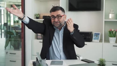 A man in a business suit is sitting at a desk in an office and arguing with someone off-camera, show
