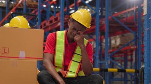 A tired black male warehouse worker sits in a paper warehouse, removes his helmet, and wipes sweat f