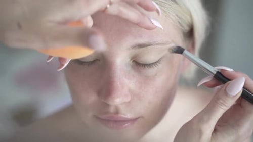 Blond women getting slowly careful eye shadow primer dispersed with sponge on natural face without m